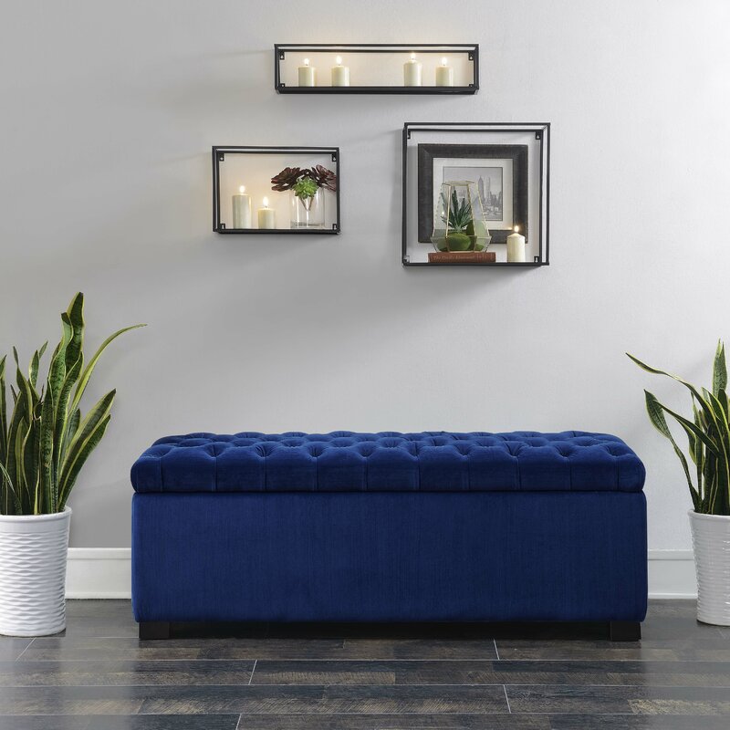 A royal blue upholstered storage bench against a grey wall with three black cube shelves holding candles and plants. 
