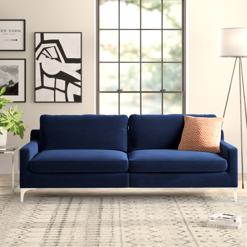 a picture of a blue couch with one accent pillow, a side table with a plant, and a stack off books upon the carpeted floor
